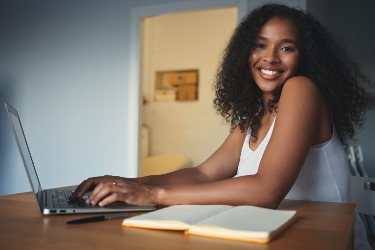 People, lifestyle, modern technology, job and communication concept. Attractive young dark skinned female freelancer with curly hair having pleased look, enjoying distant work, using generic laptop